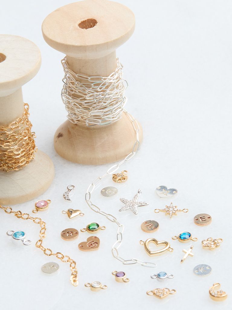 Gold and silver permanent jewelry chains on wooden spools beside an assortment of charms